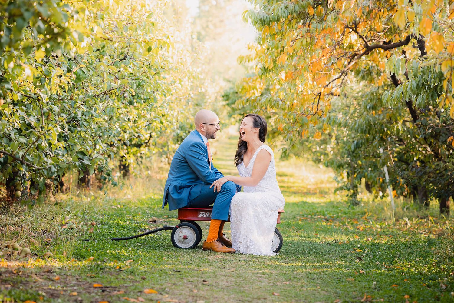 Playful wedding photo with couple sitting on small red wagon in autumn orchard.
