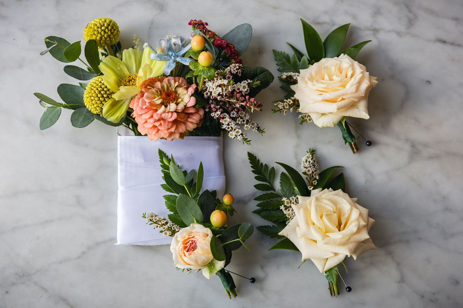 Wedding boutonnieres and corsages with peach roses, yellow craspedia, and greenery arranged on marble surface.