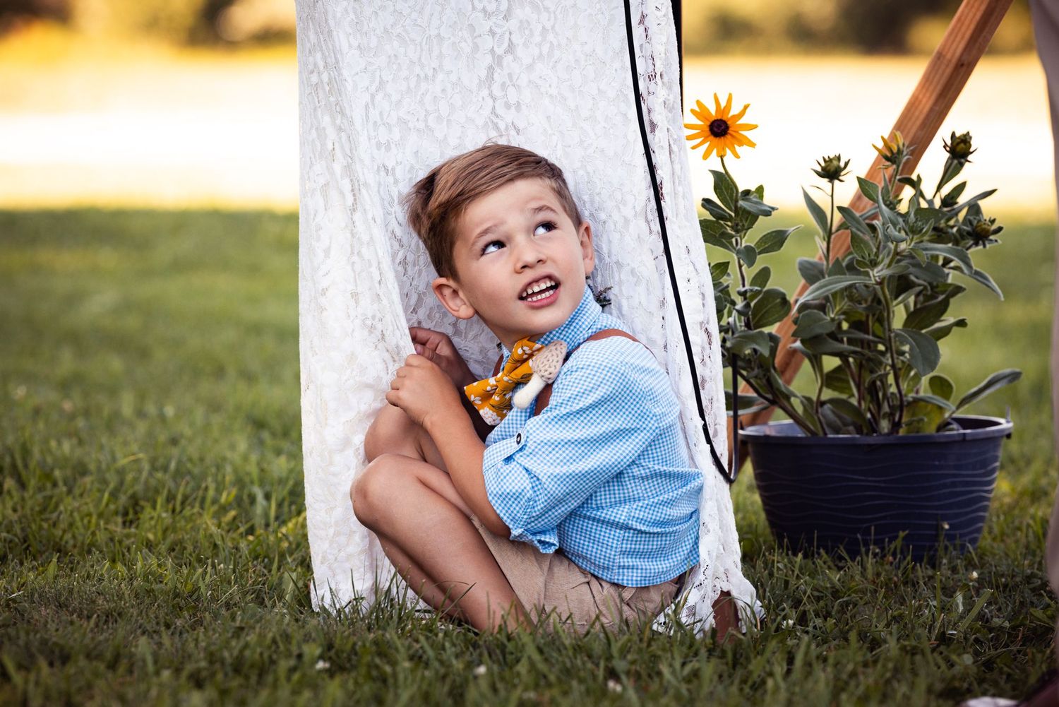 A young child in a blue shirt sits beside a sunflower plant in a garden on a sunny summer day.