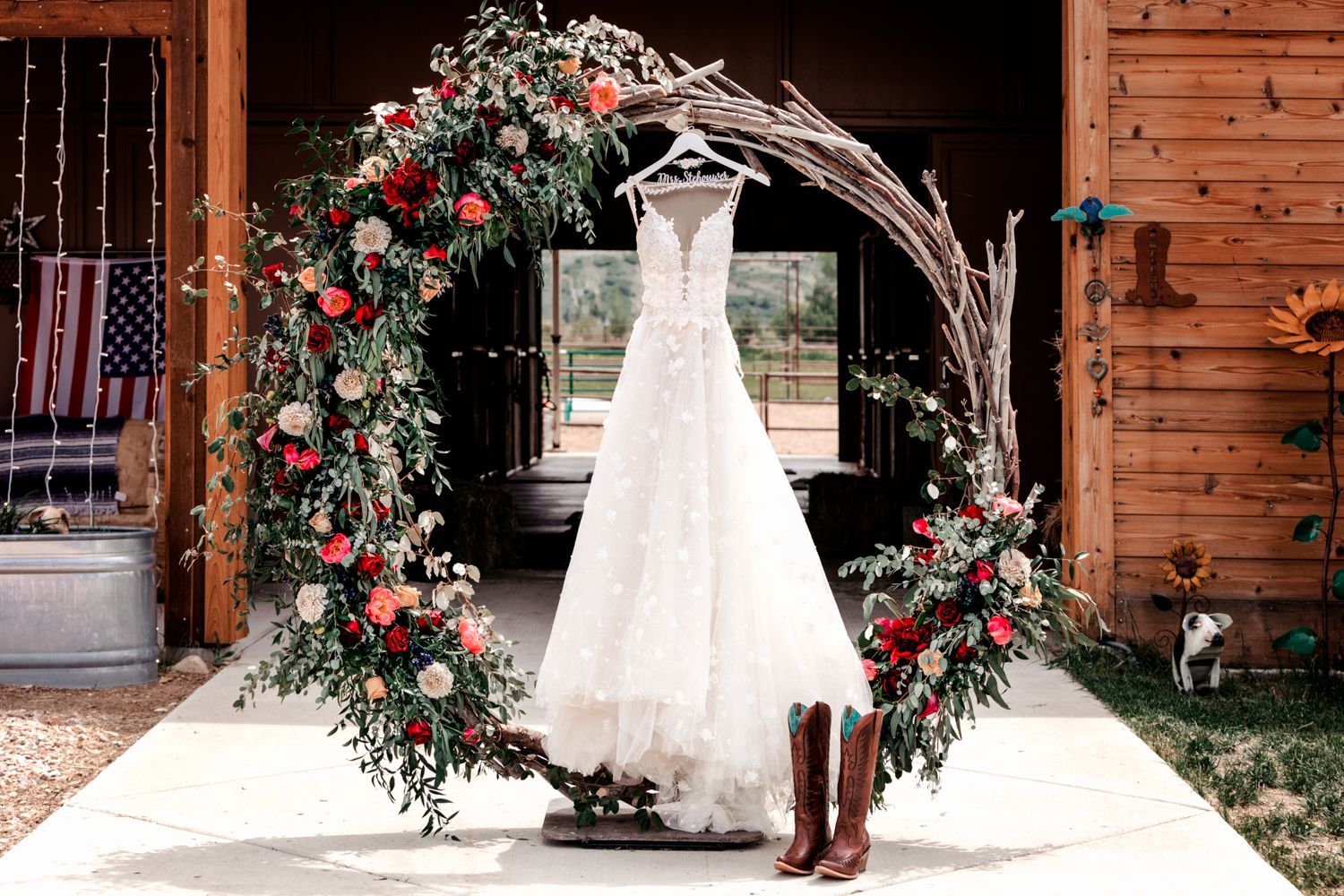 A white lace wedding dress hangs on a circular floral arch decorated with red roses and greenery, with brown boots below.
