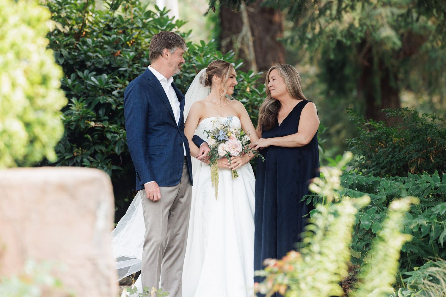 Bride in white gown with bouquet stands between groom in navy blazer and officiant in navy dress during outdoor wedding ceremony.