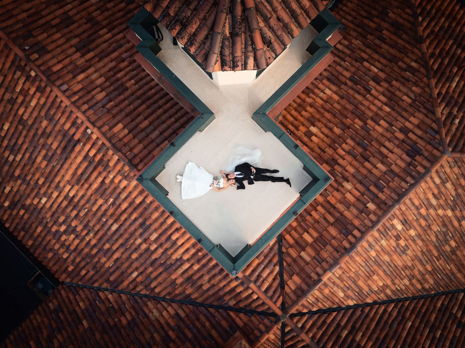 Aerial view of bride and groom lying on terracotta roof tiles in a diamond-shaped skylight during wedding photography.
