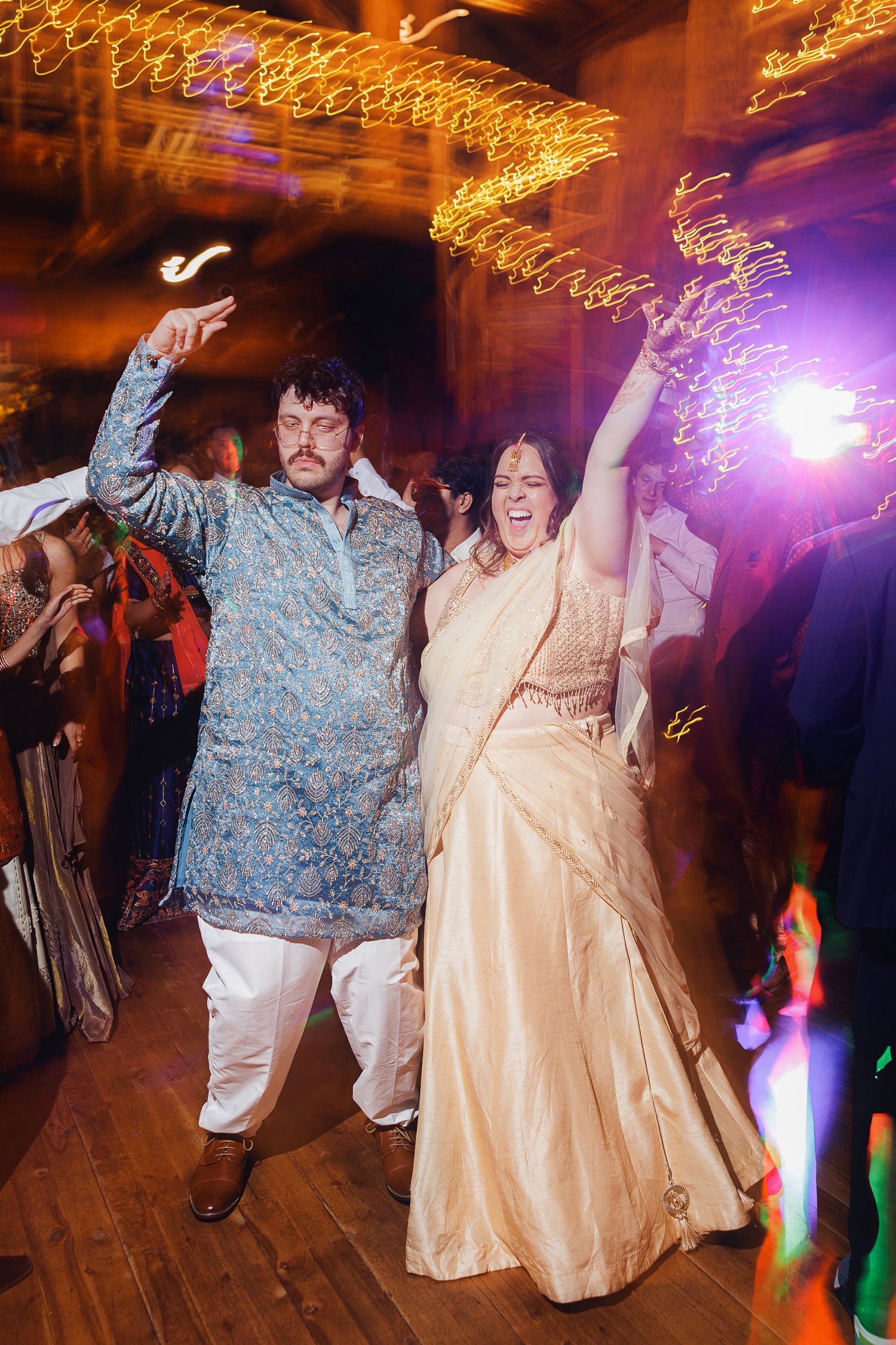 Couple in traditional Indian attire dancing joyfully at a wedding reception with sparkling lights.