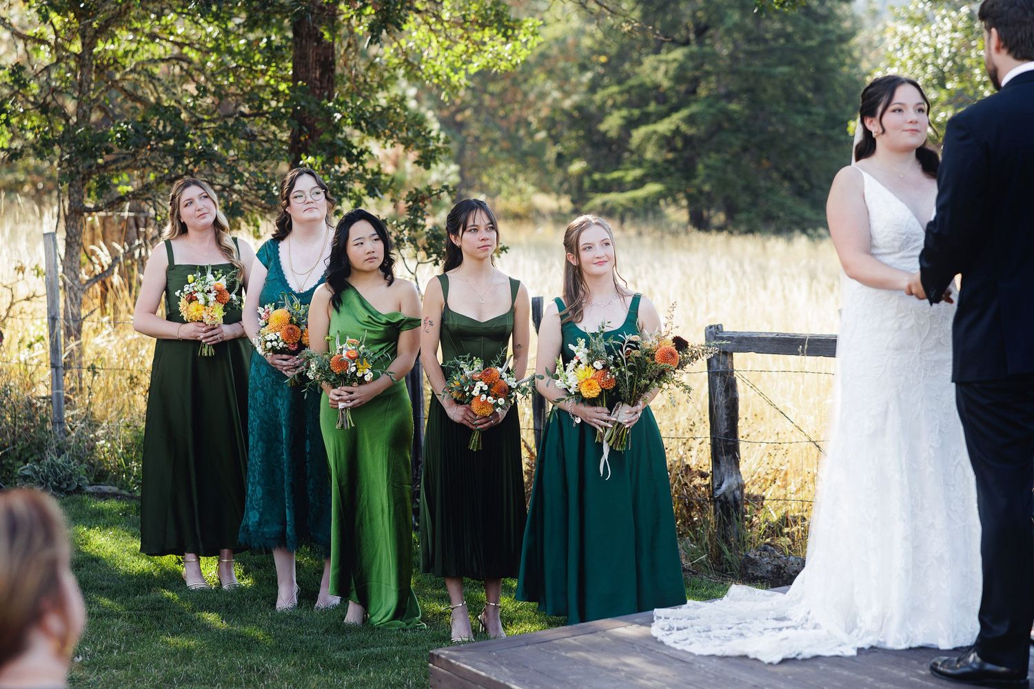Bridesmaids in green dresses holding colorful bouquets stand beside a bride in white at an outdoor wedding ceremony.