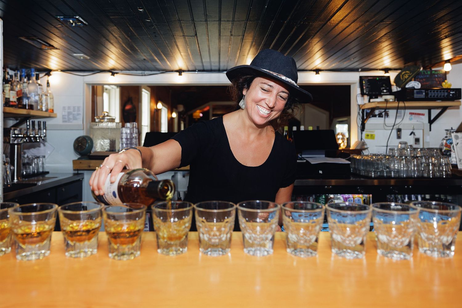 Bartender in black hat pouring whiskey into a row of shot glasses on a wooden bar counter.