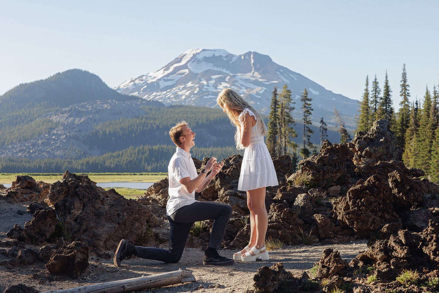 Man proposes to woman on one knee near volcanic rocks with snow-capped mountain backdrop in Oregon.