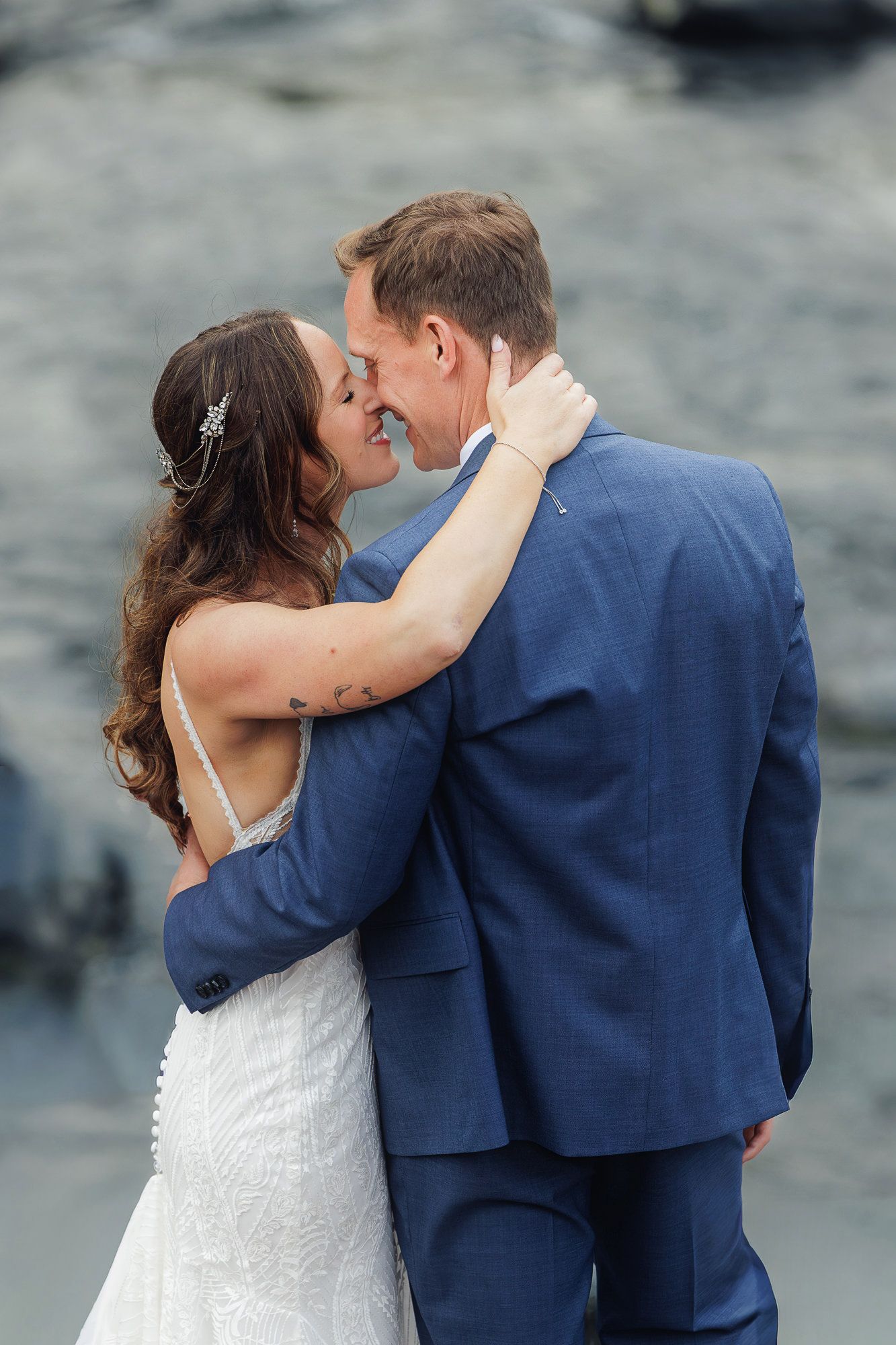A romantic wedding couple embraces against rocky coastal backdrop, bride in white lace and groom in navy blue suit.