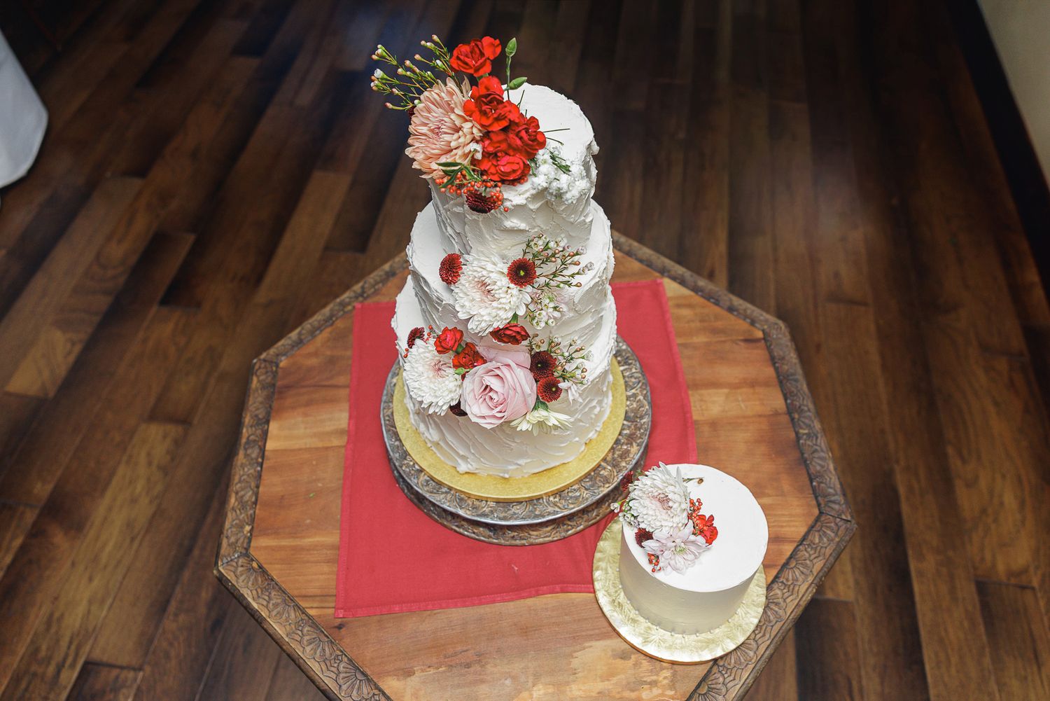 Wedding cake decorated with red and pink roses sits on wooden table with multiple tiers and elegant design.
