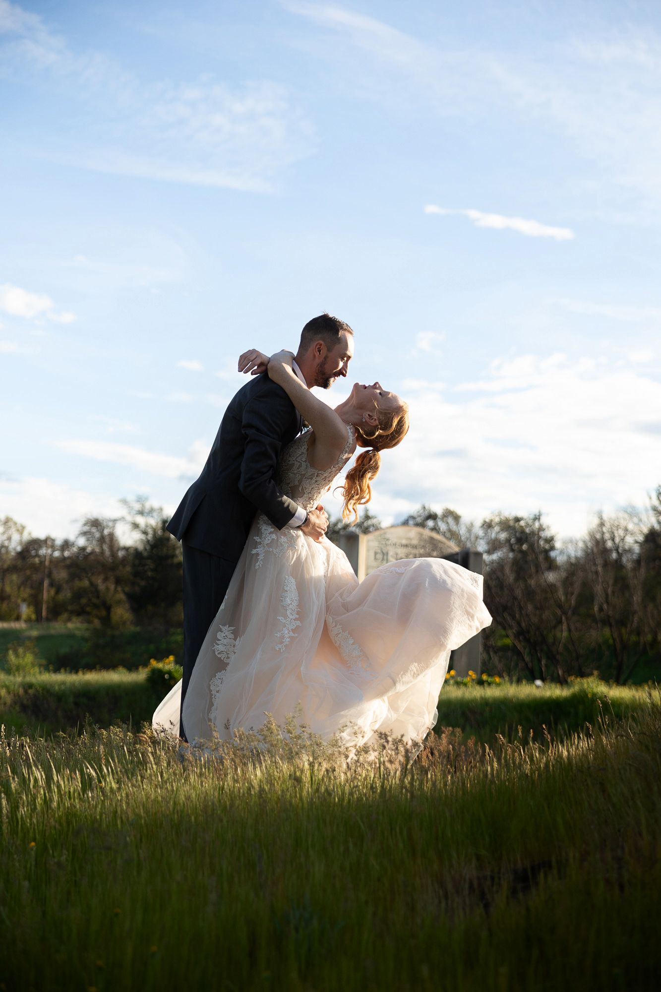 Bride and groom share a romantic dip kiss in a lush green field at golden hour on their wedding day.