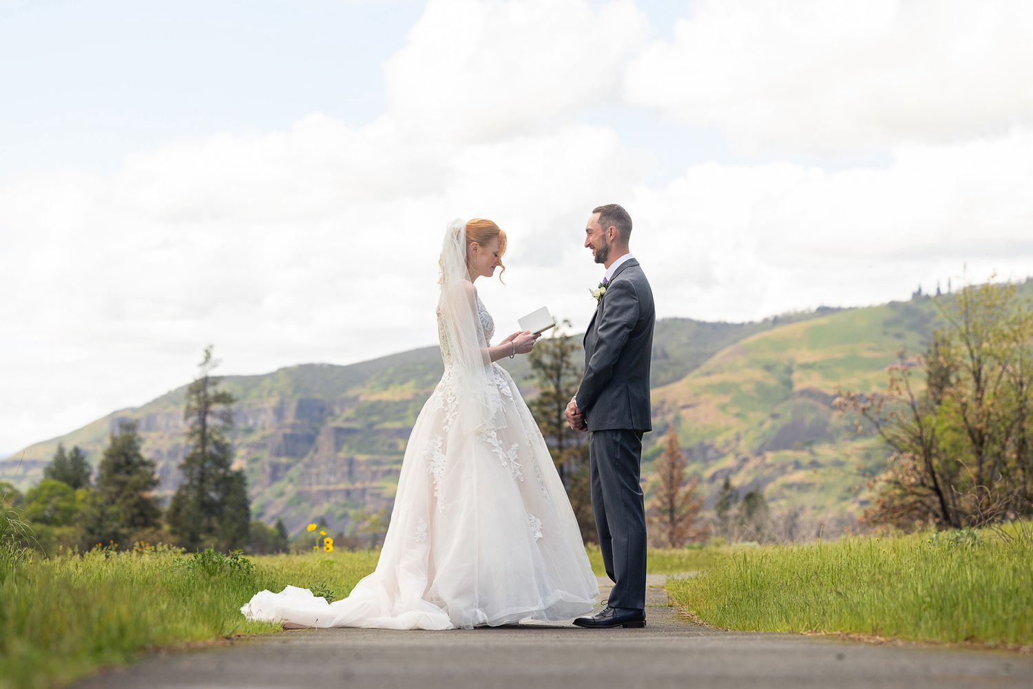 Bride and groom stand facing each other on a path with lush green hills and cloudy sky backdrop.