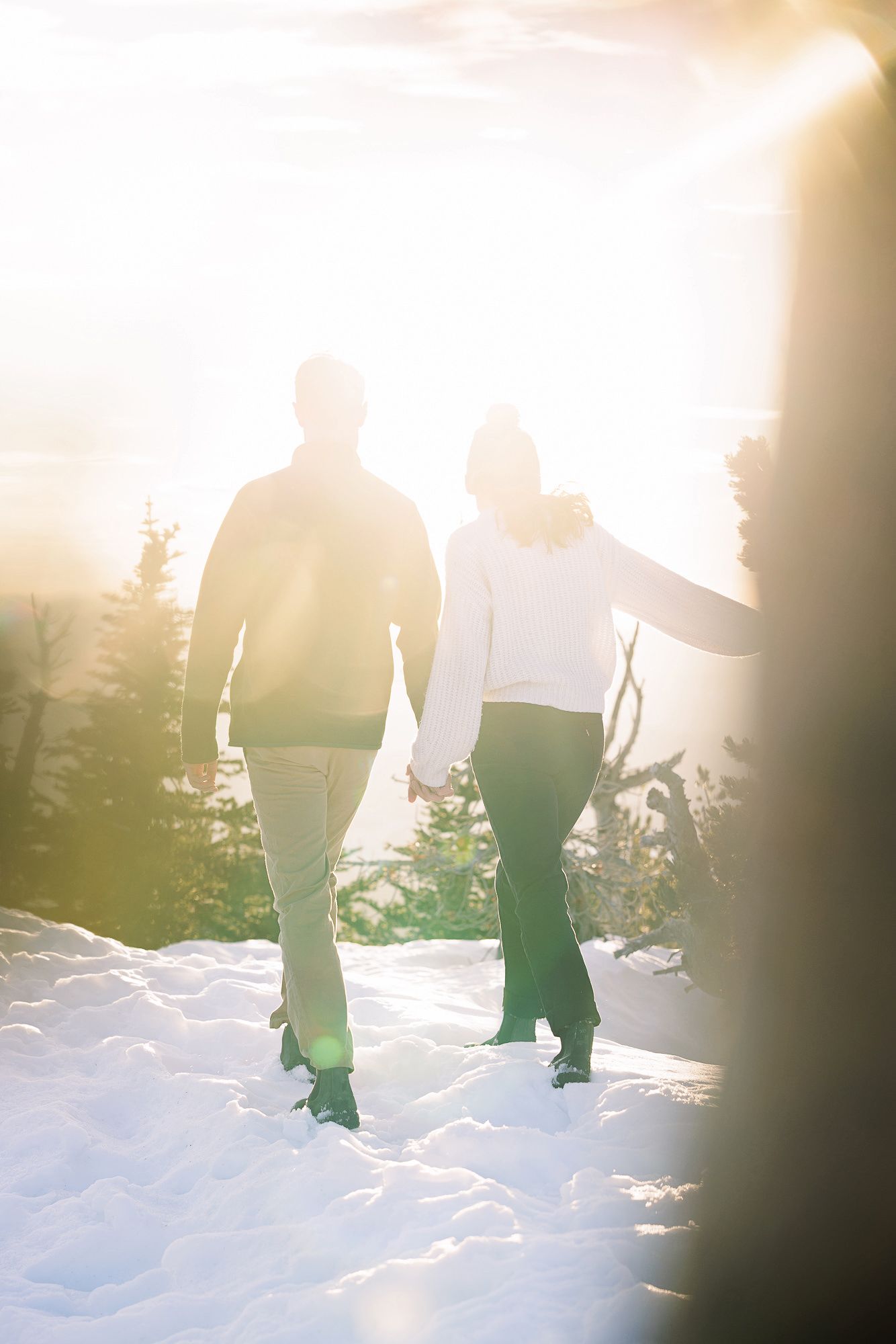 Couple holding hands walking through snowy winter landscape with bright sunlight and pine trees in background.
