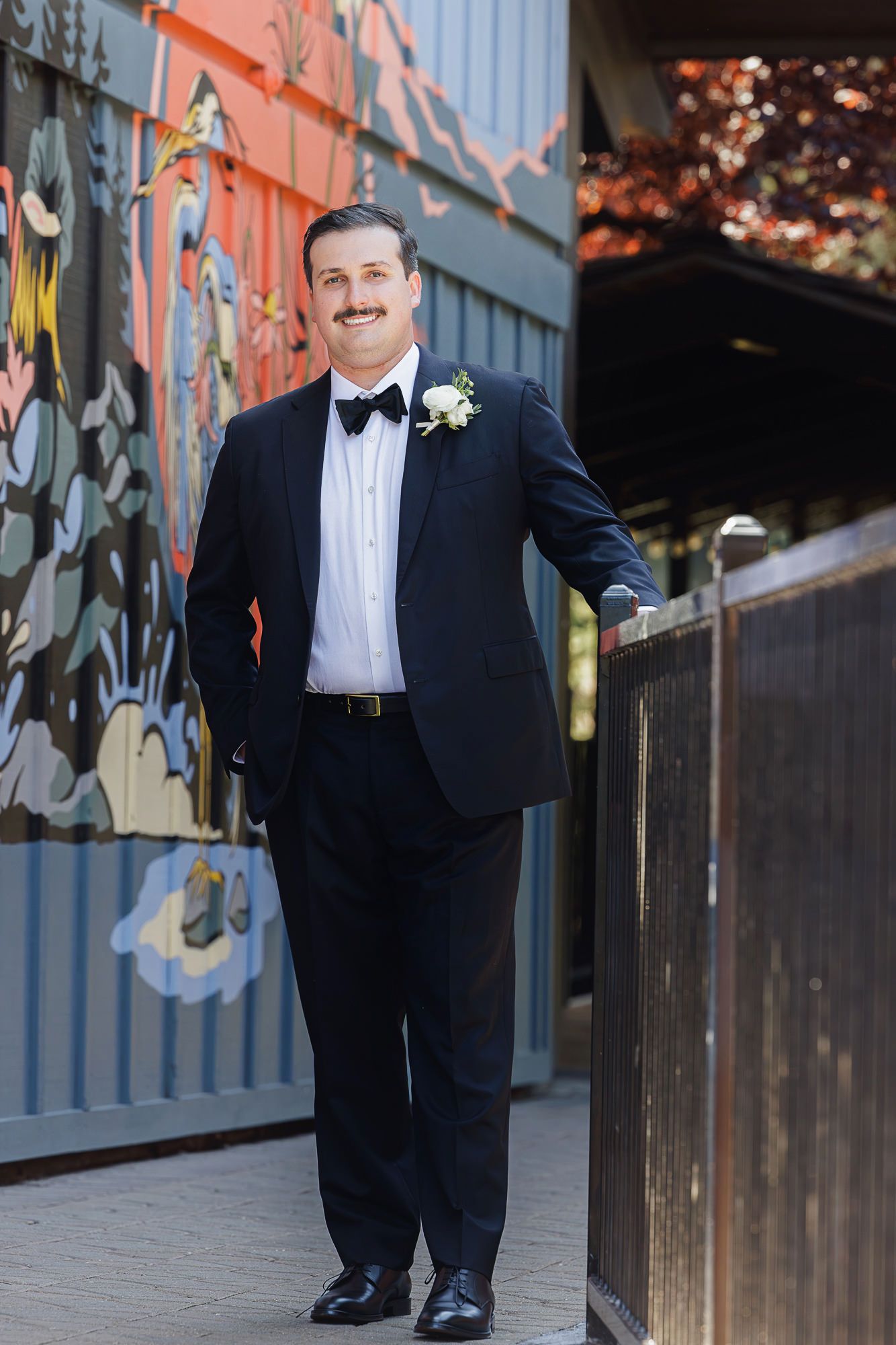 Smiling groom in navy tuxedo with bow tie and white boutonniere poses beside colorful mural wall.