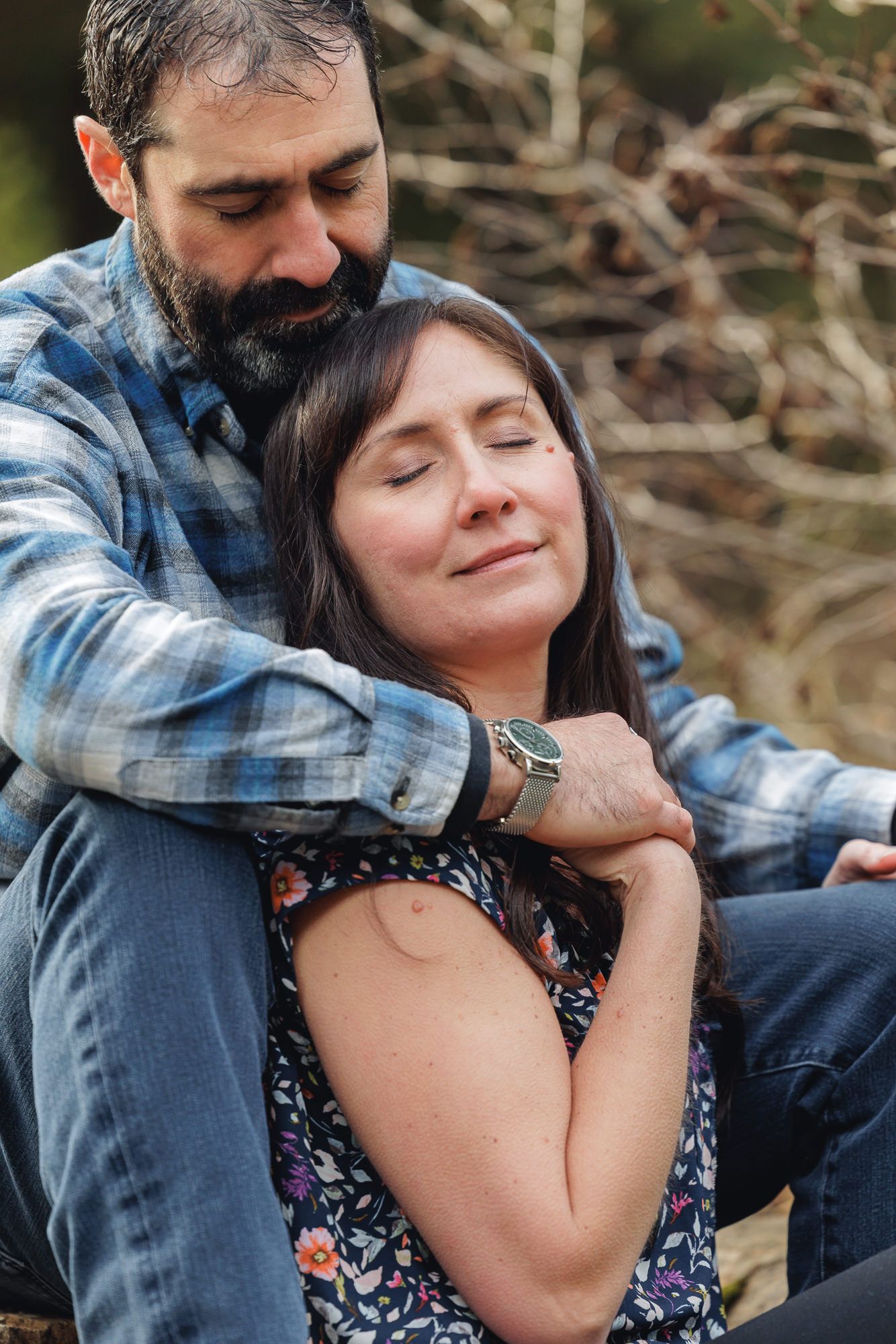A bearded man embraces a woman with dark hair from behind, both sitting outdoors in a tender, peaceful moment.