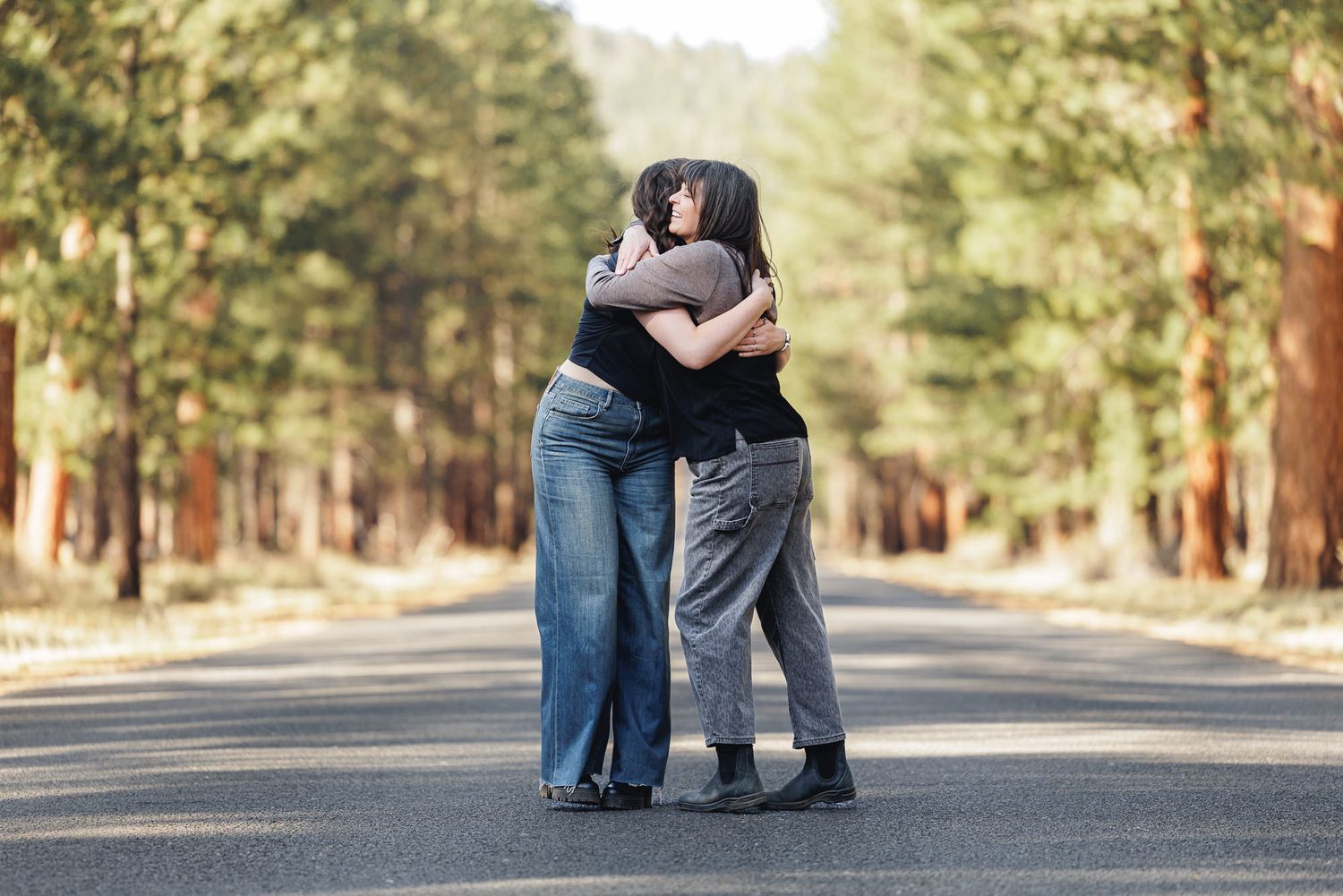 Two women sharing a warm embrace on a tree-lined road surrounded by tall pine trees.