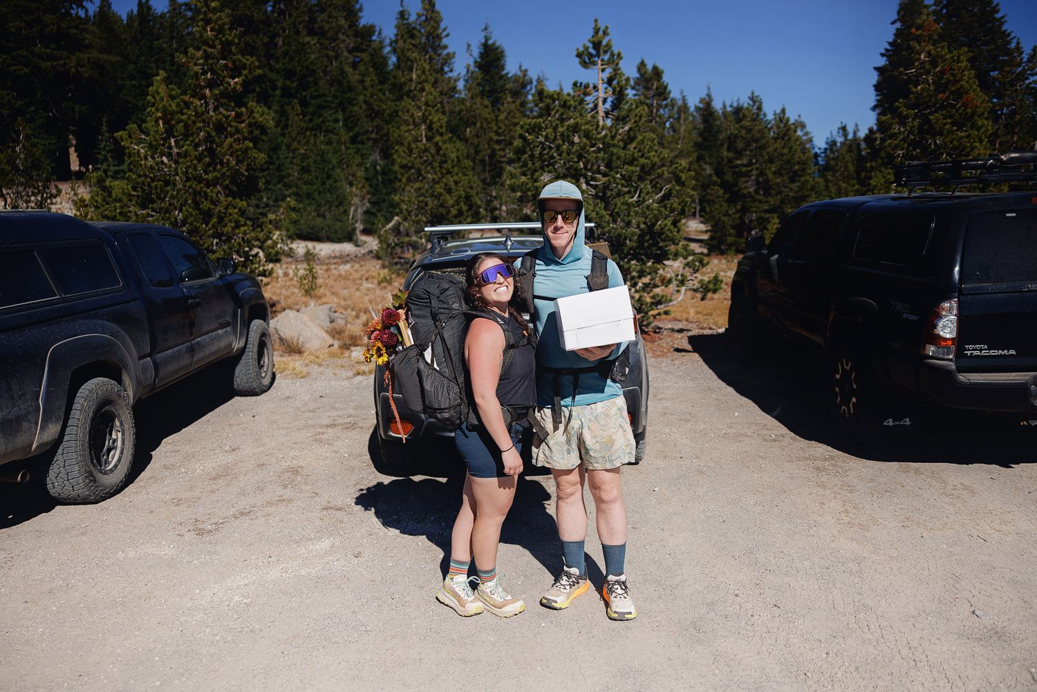 Two hikers with backpacks pose together holding a paper in a dirt parking area beside parked vehicles, with evergreen trees behind them.