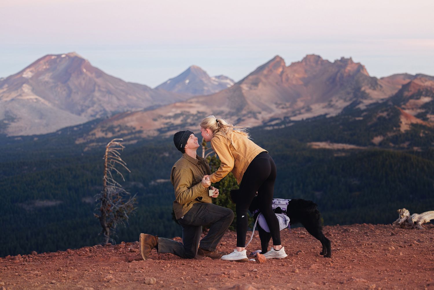 Man kneeling on one knee proposing to a woman on a mountain summit at sunset, with a black dog beside them and rugged peaks behind.