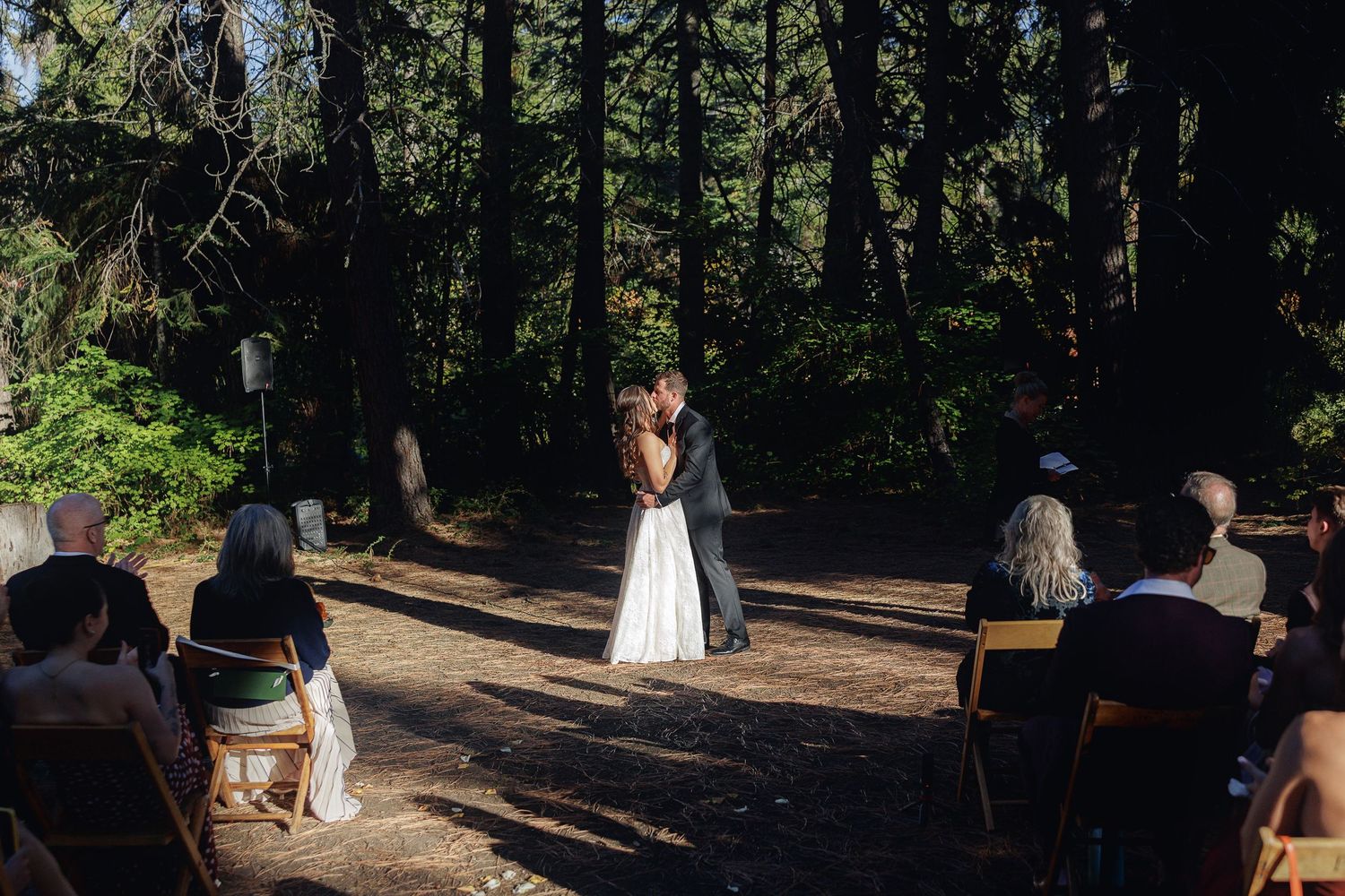 Bride and groom share first kiss at outdoor forest wedding ceremony with guests seated on wooden chairs.