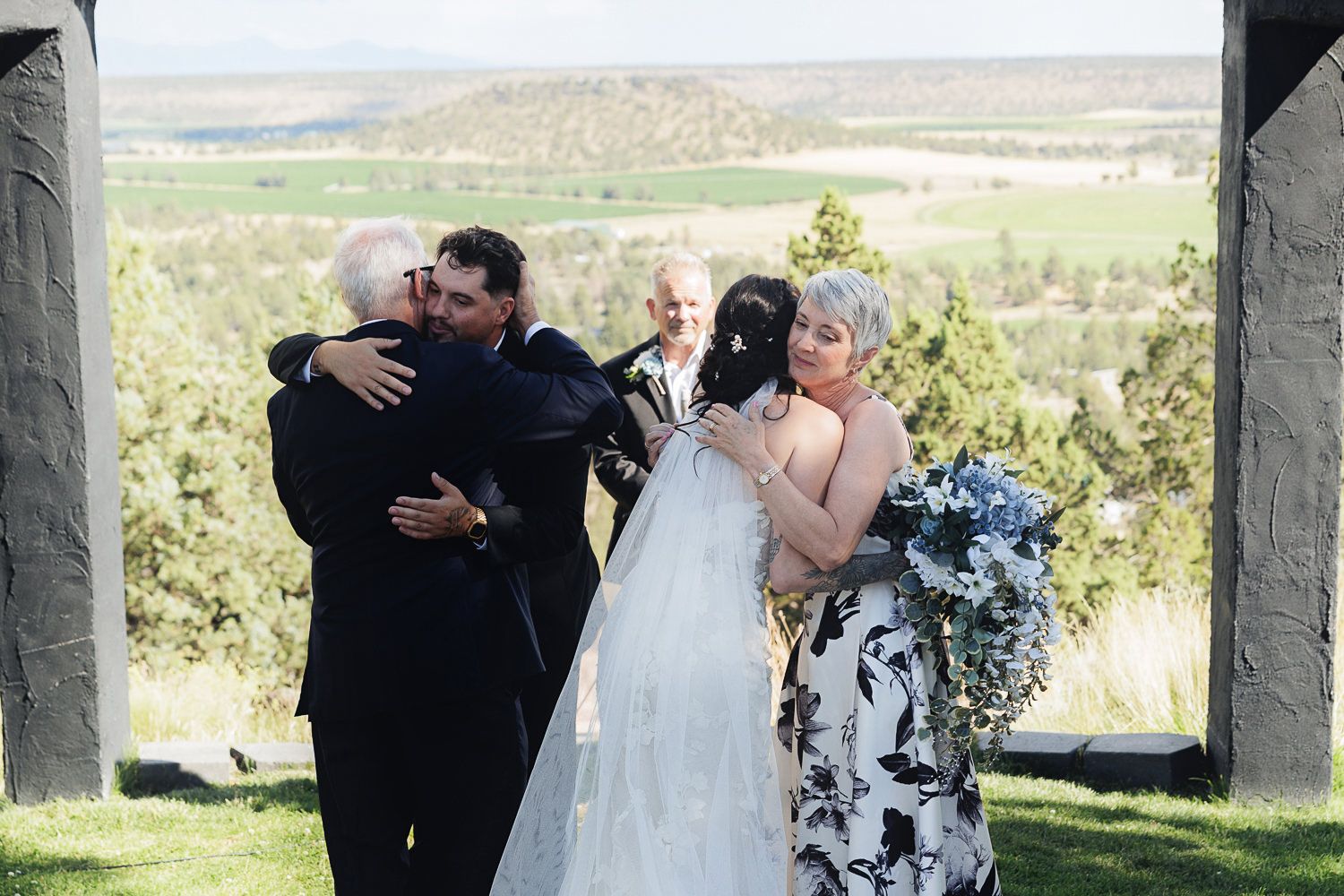 Wedding party members embrace in warm greeting outdoors with rolling green countryside landscape in the background.