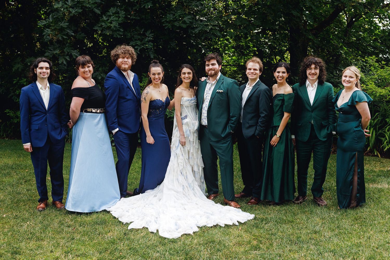 Wedding party poses outdoors in formal attire, bride in white gown flanked by guests in navy and emerald green dresses.