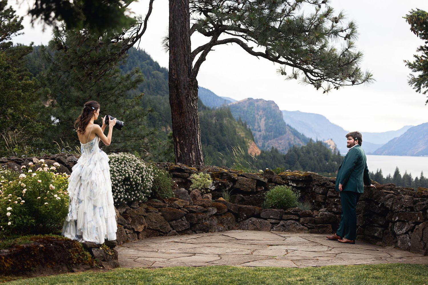 Bride photographs groom in teal suit at scenic mountain overlook with stone wall and pine tree backdrop.