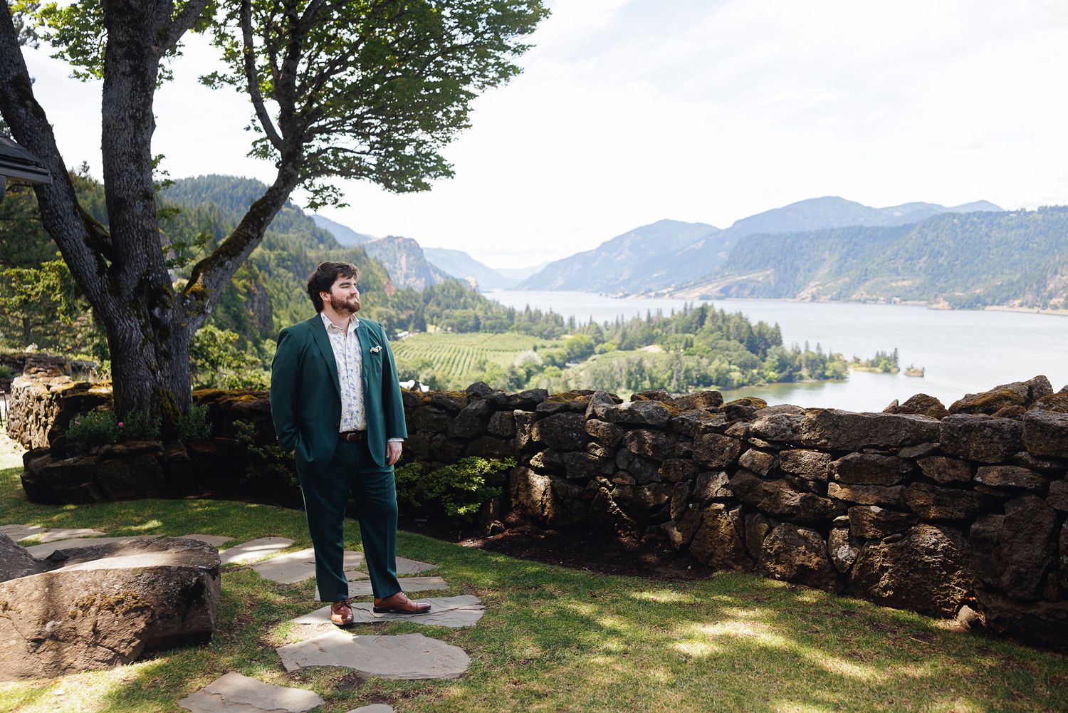Man in green suit standing on stone terrace overlooking scenic river valley and mountains on sunny day.