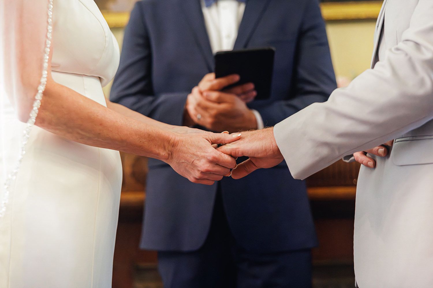 Close up of hands exchanging rings during wedding ceremony as officiant holds phone in background.