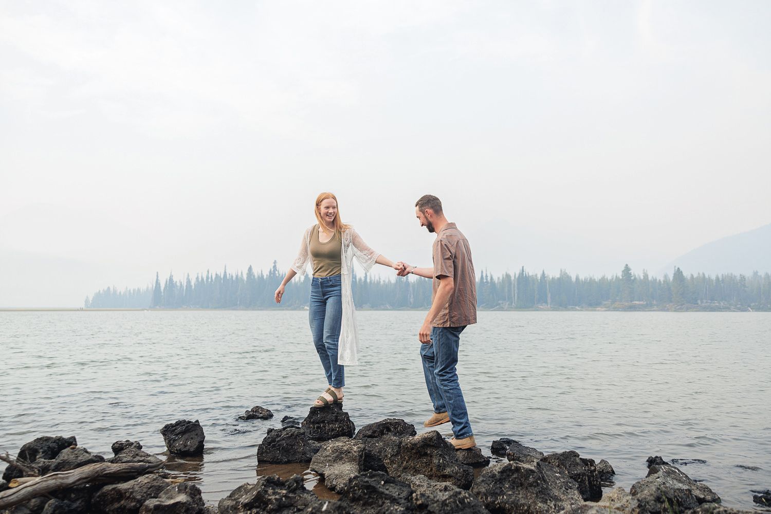 Couple walks hand in hand on dark rocky beach shoreline with calm ocean water and cloudy sky in background.