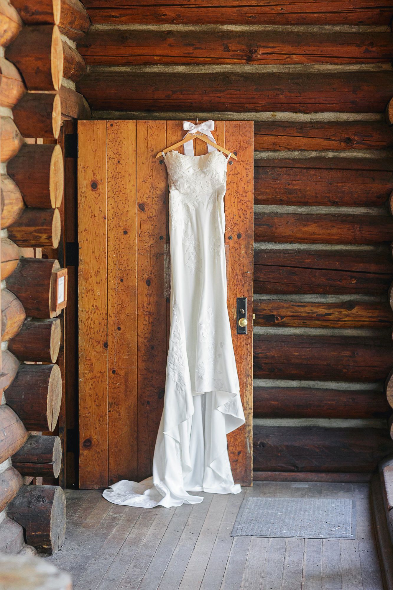 A white vintage christening gown hangs on a wooden wall in a rustic log cabin setting.
