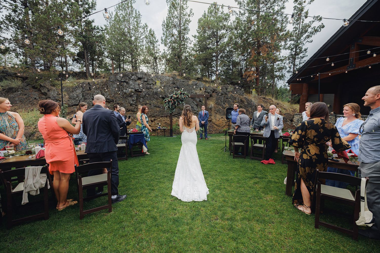 Wedding guests gather under string lights and tall pine trees at an outdoor reception venue during sunset.