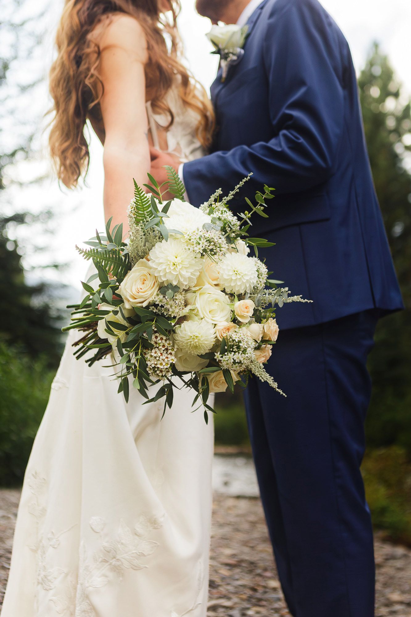 A couple in navy and white wedding attire holds a romantic white and cream floral bouquet during their outdoor ceremony.