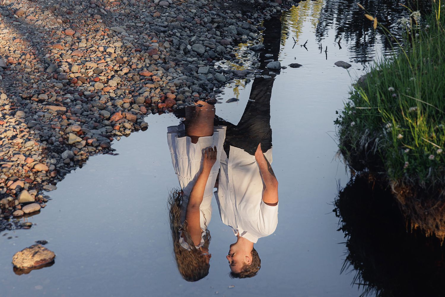 A person's reflection captured upside down in a calm shallow water puddle surrounded by rocks and green grass.