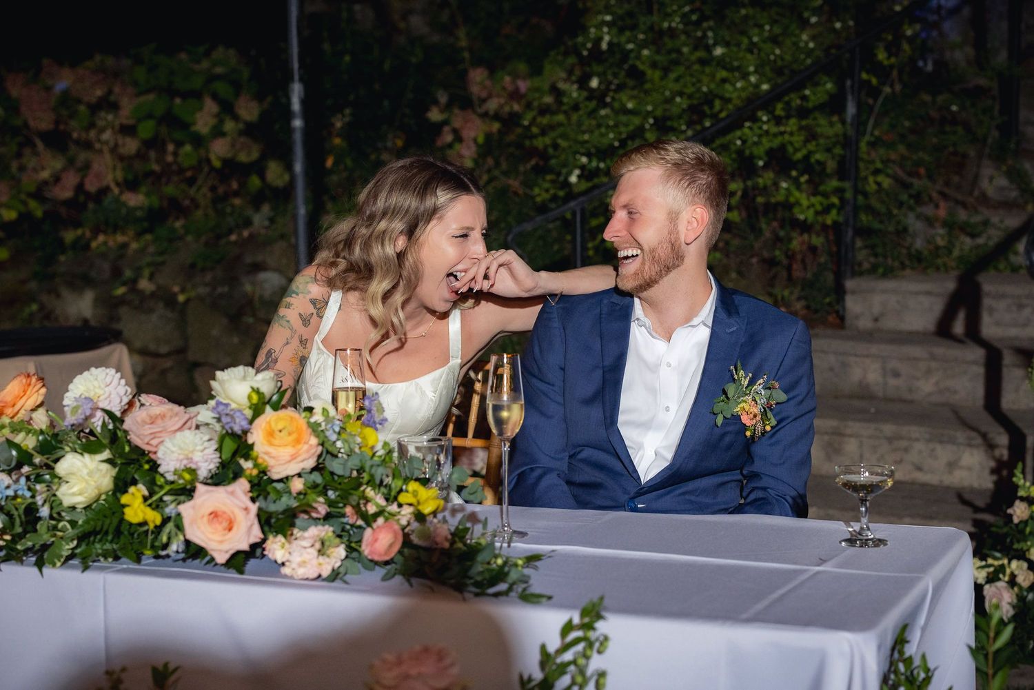 A couple in wedding attire share a sweet moment at their reception table with colorful floral arrangements and wine glasses.