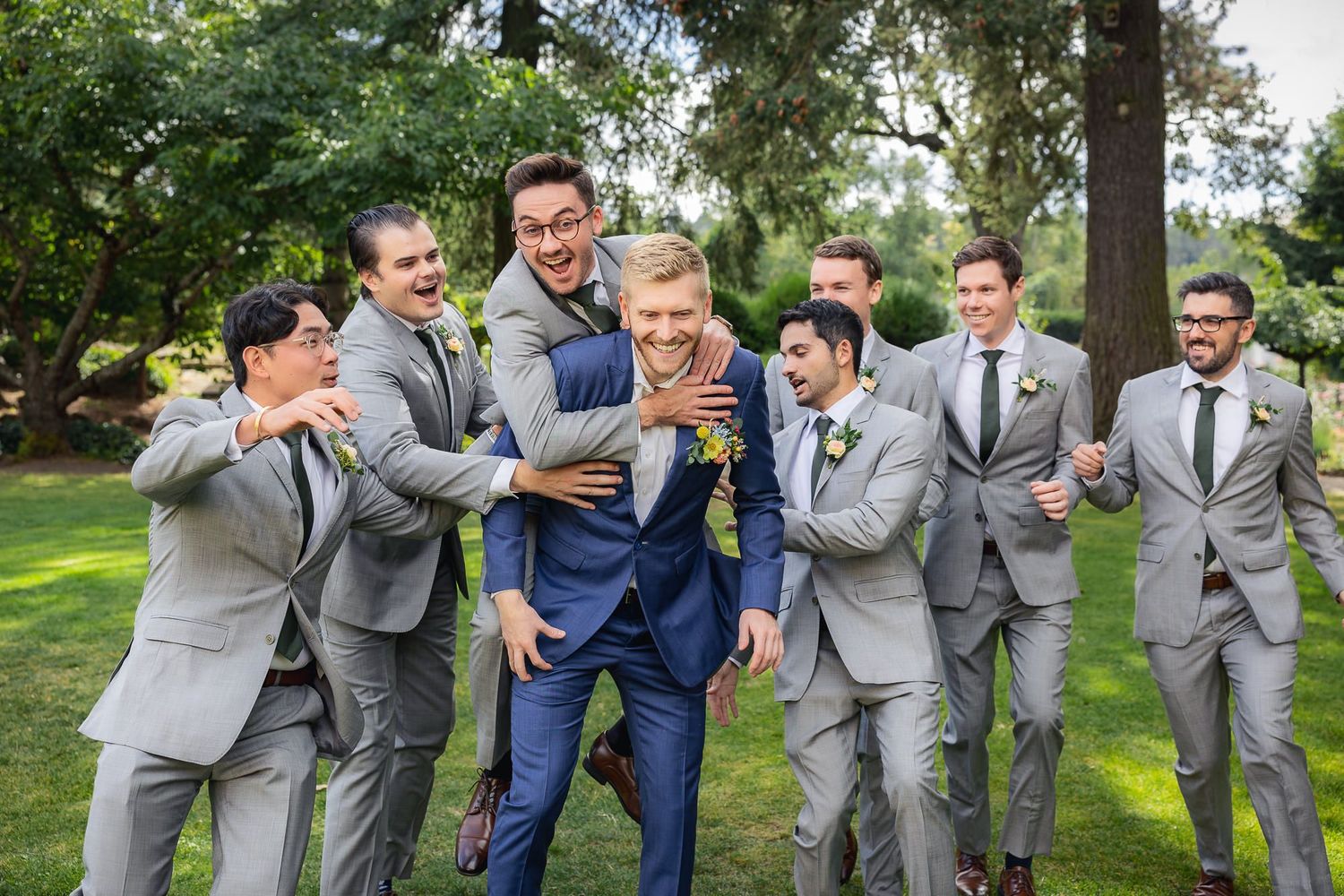 Wedding party in matching gray suits laughs and celebrates together during outdoor photo session at sunny park.