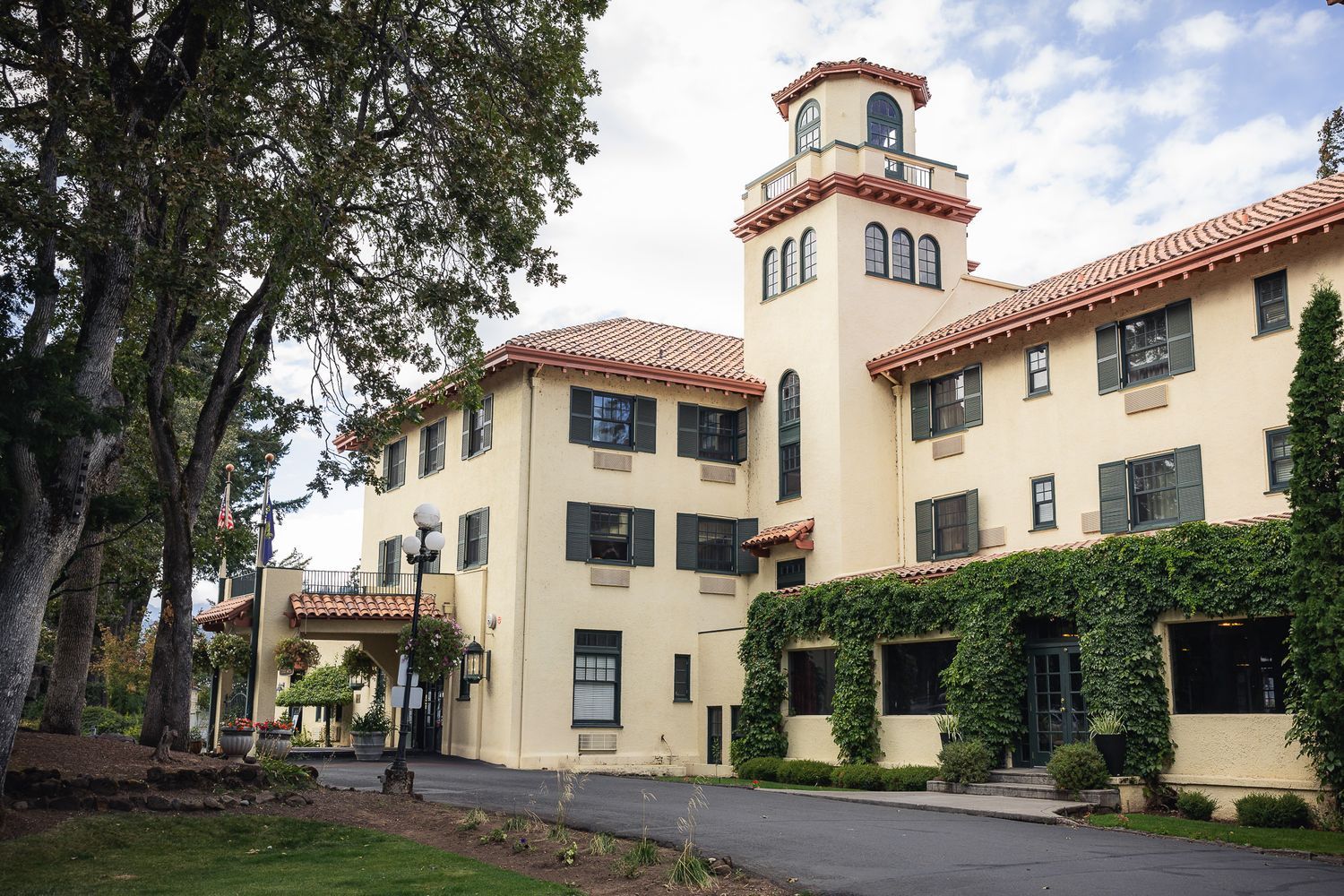 Historic Spanish-style building with cream walls, tower and red tile roof on a university campus surrounded by green trees.