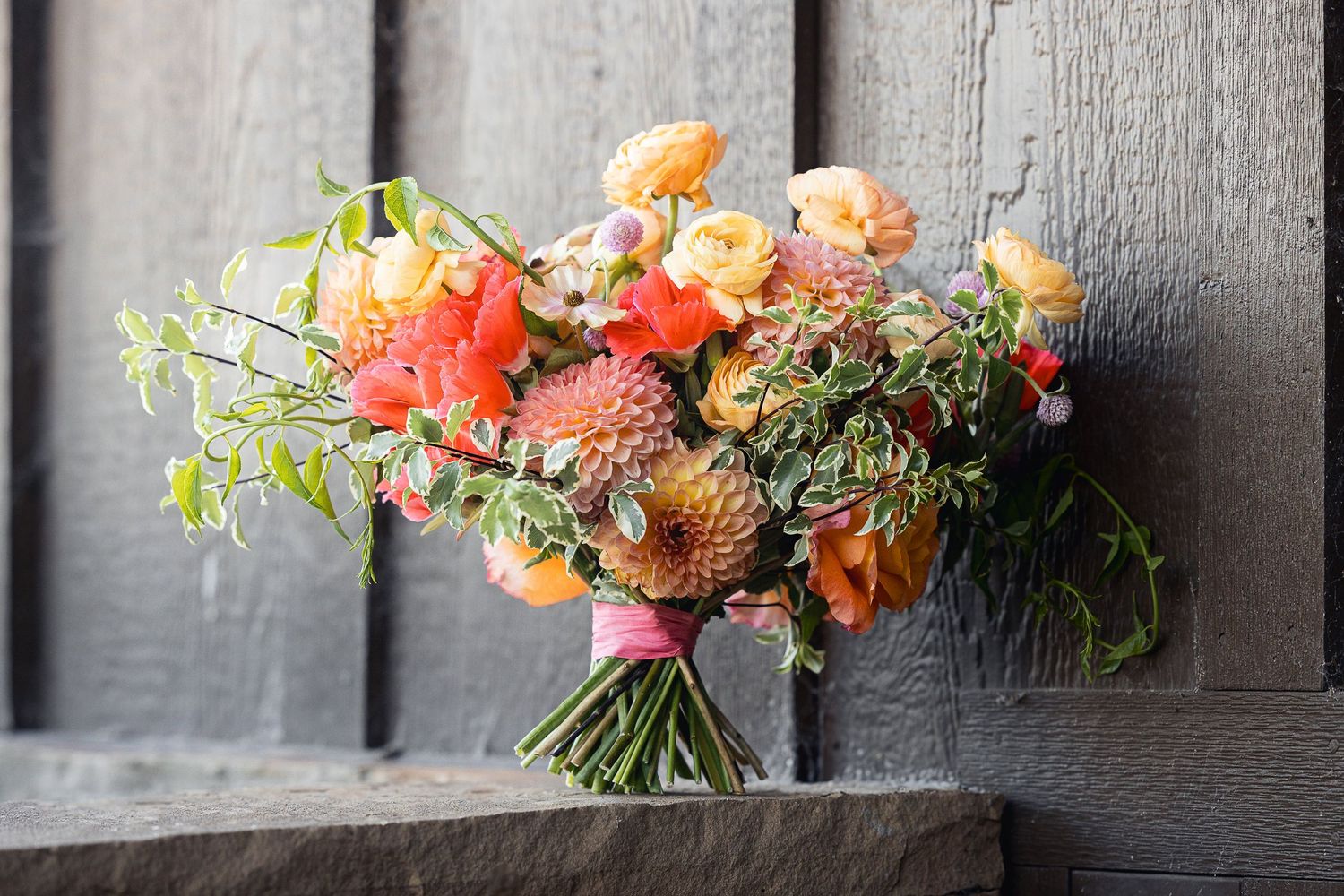 Vibrant coral and peach floral arrangement with greenery against a grey wooden background.