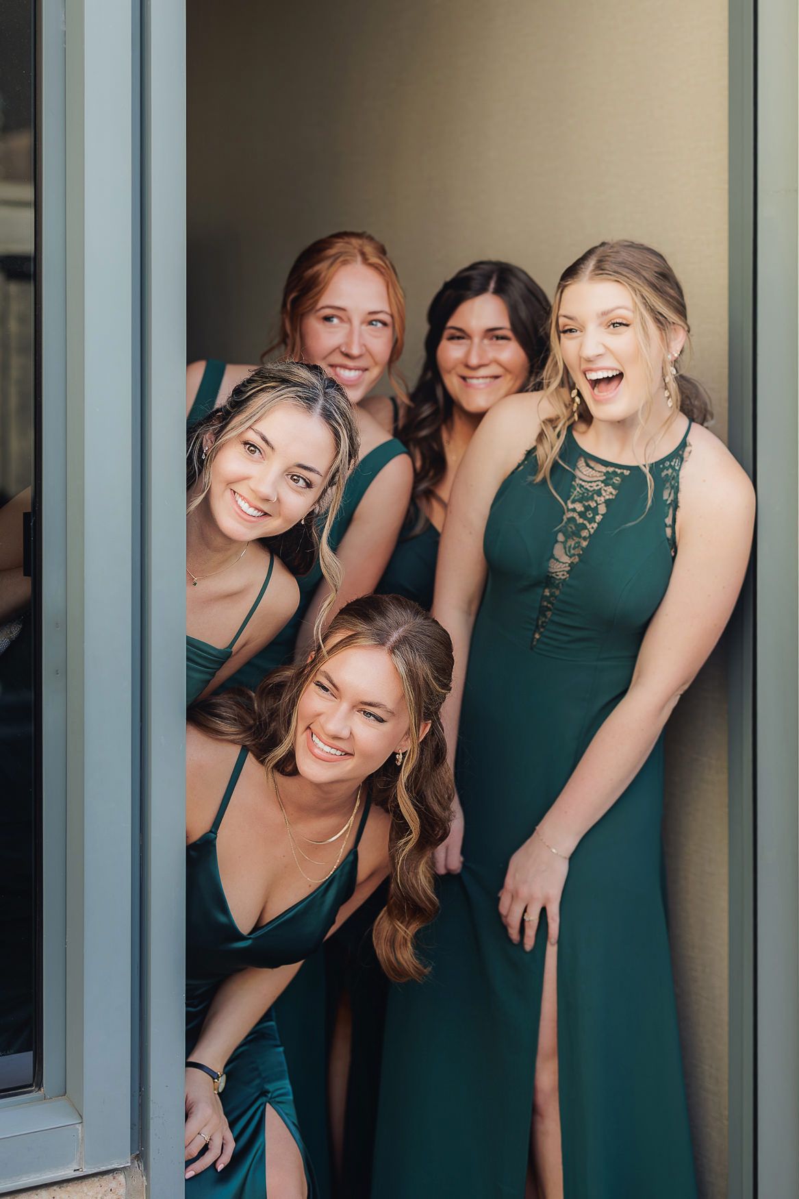 Bridesmaids in emerald green dresses pose together playfully in a doorway.