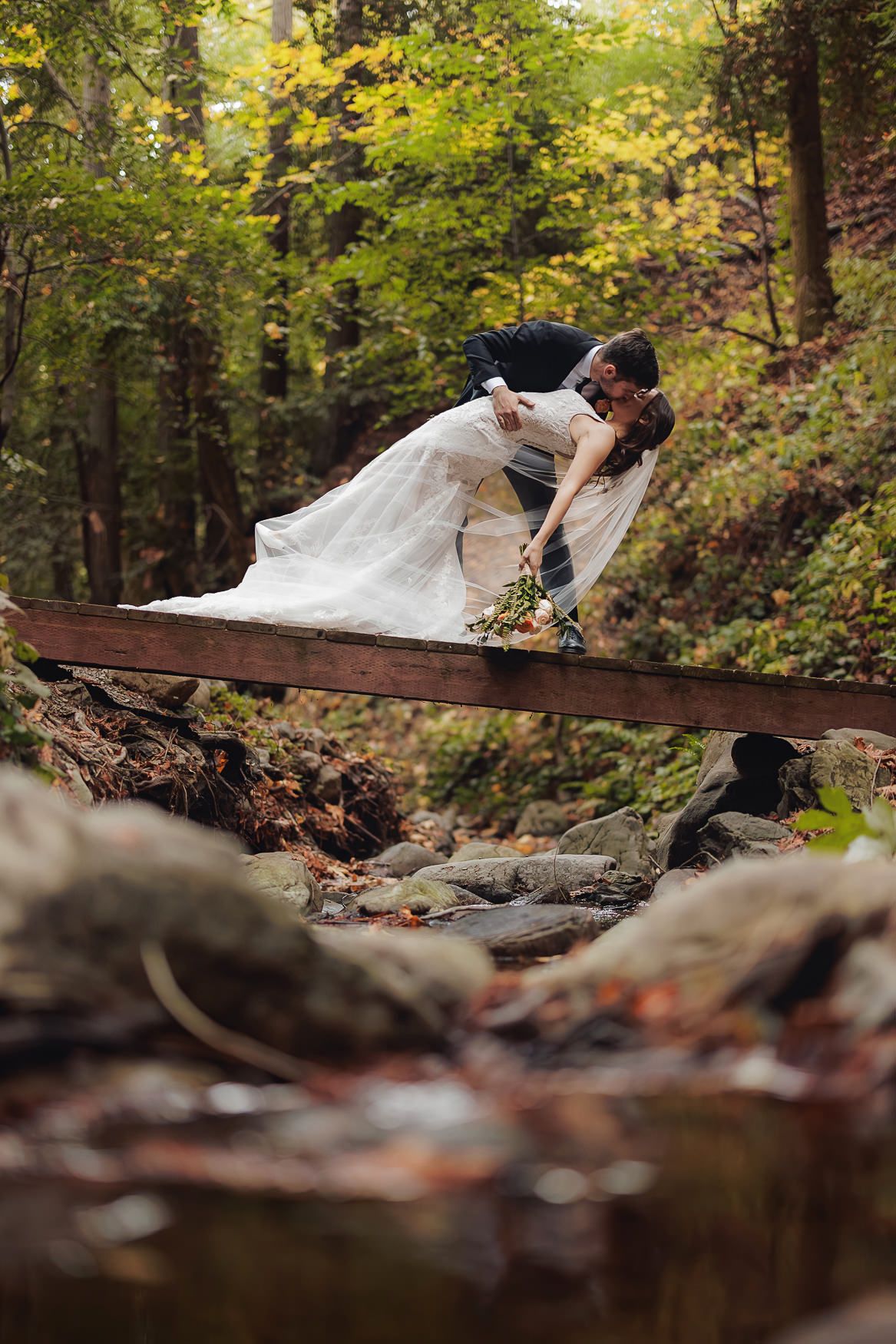 A bride in a flowing white dress lounges on a rustic wooden footbridge over a stream in an autumn forest setting.
