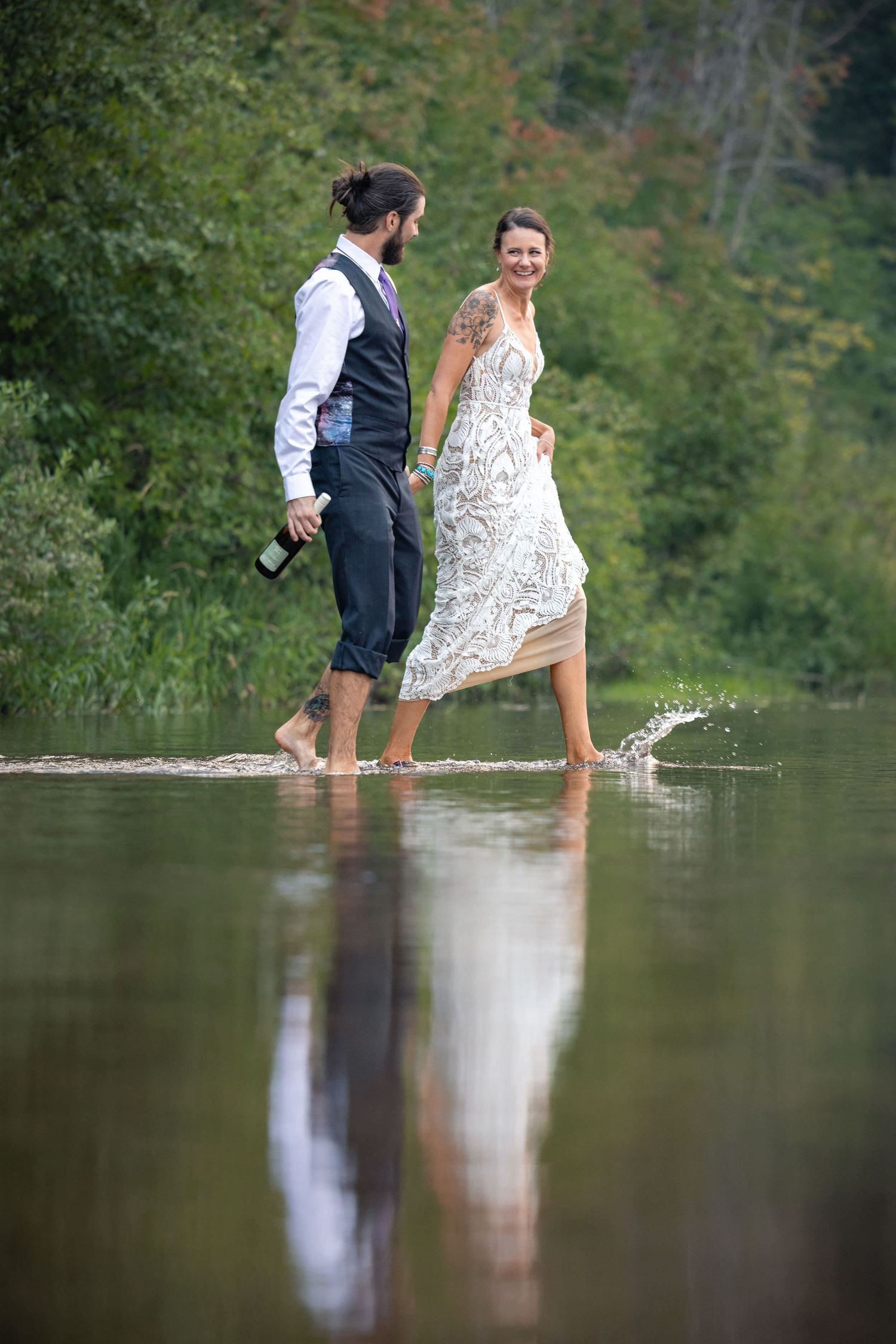 A couple splashes and plays in shallow water, with the person in a white lace dress and other in formal vest attire.