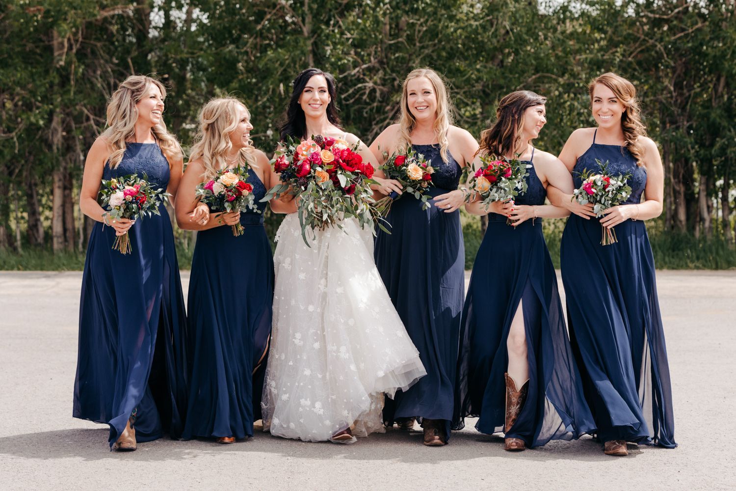 Bridal party in flowing navy blue dresses poses outdoors with the bride in white holding vibrant red flowers.