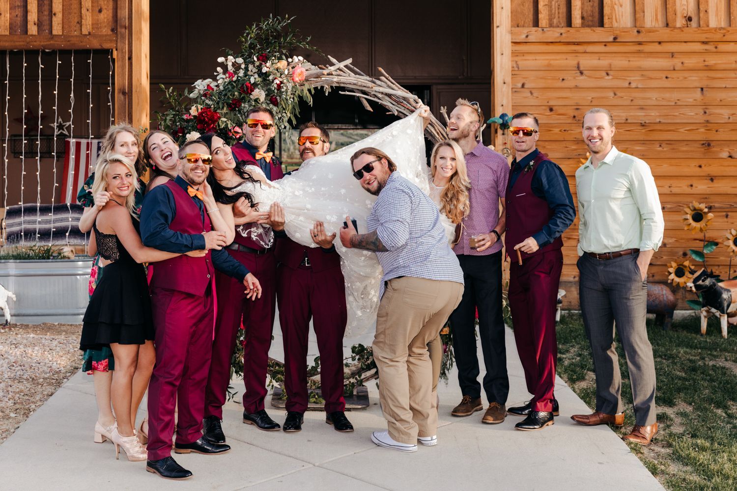 A wedding party poses with fun energy and burgundy attire outside a rustic wooden barn.
