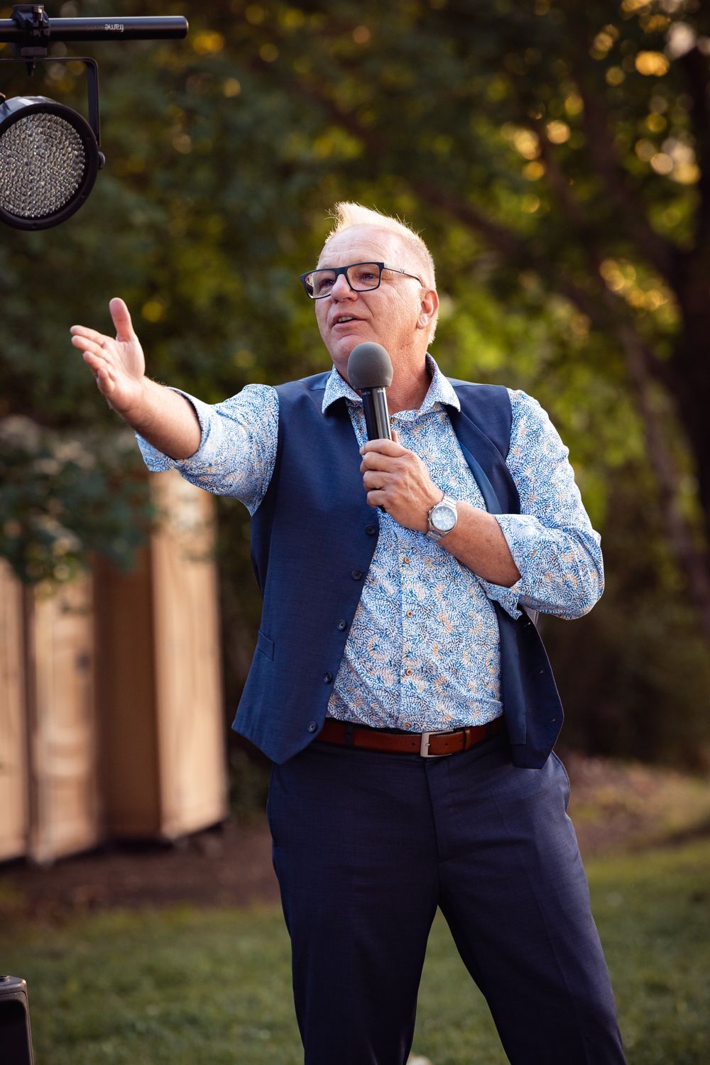A speaker wearing a navy vest and patterned blue shirt gestures while holding a microphone at an outdoor event.