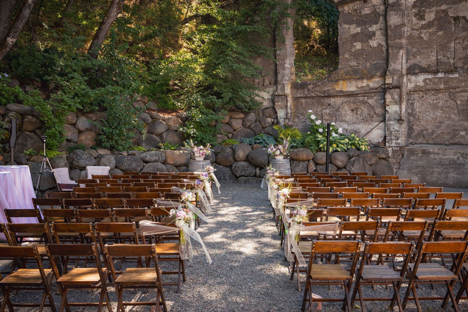 Rustic outdoor wedding ceremony setup with wooden chairs and stone wall backdrop in wooded setting.