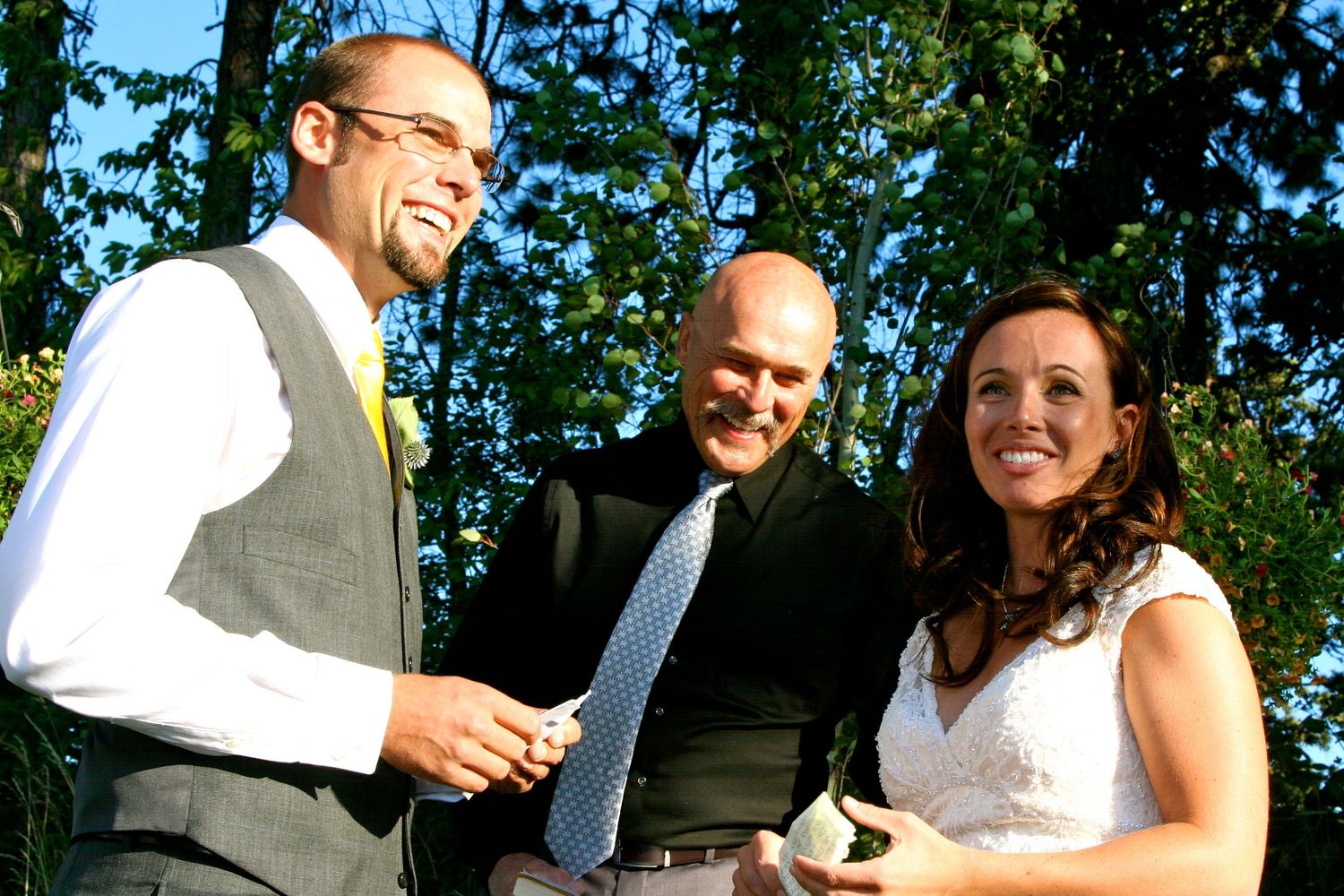 Three people at an outdoor wedding ceremony sharing a happy moment while handling a cake knife.