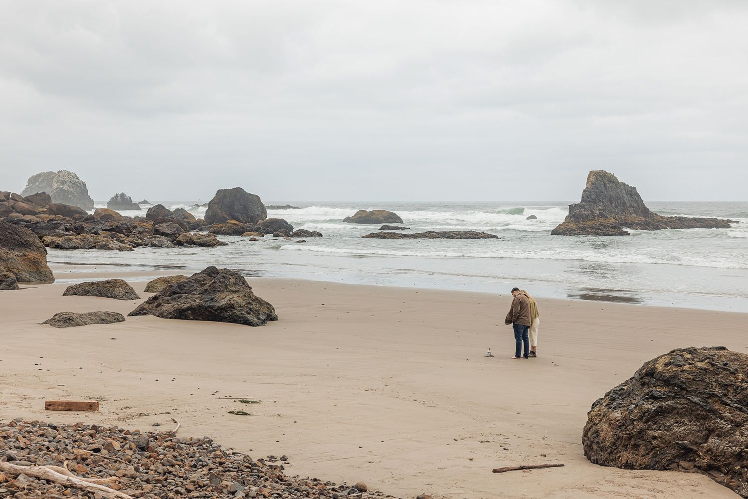 Two people stand on a sandy beach surrounded by large rocks with ocean waves and overcast skies in the background.