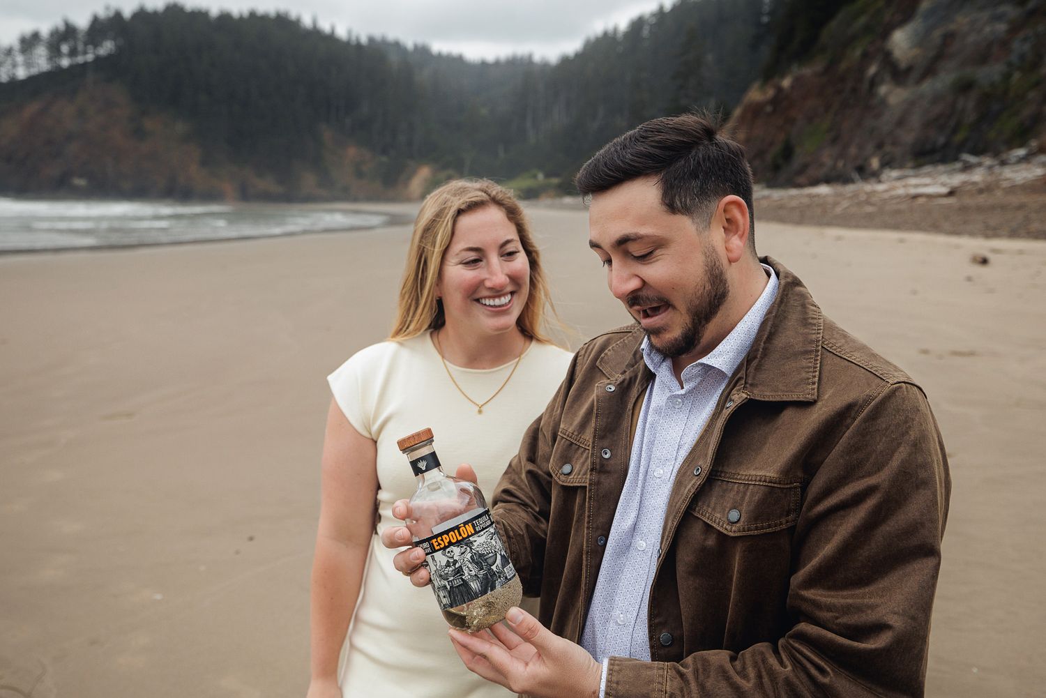 Couple smiling on a beach holding a product bottle, with ocean cliffs in the background.