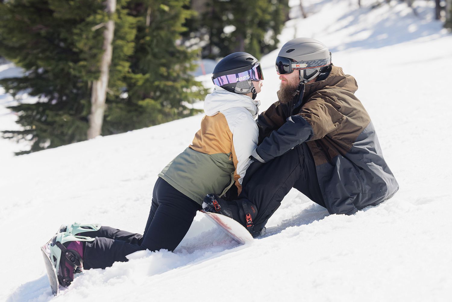 A snowboarder in winter gear sits on the snowy ground, surrounded by pine trees.