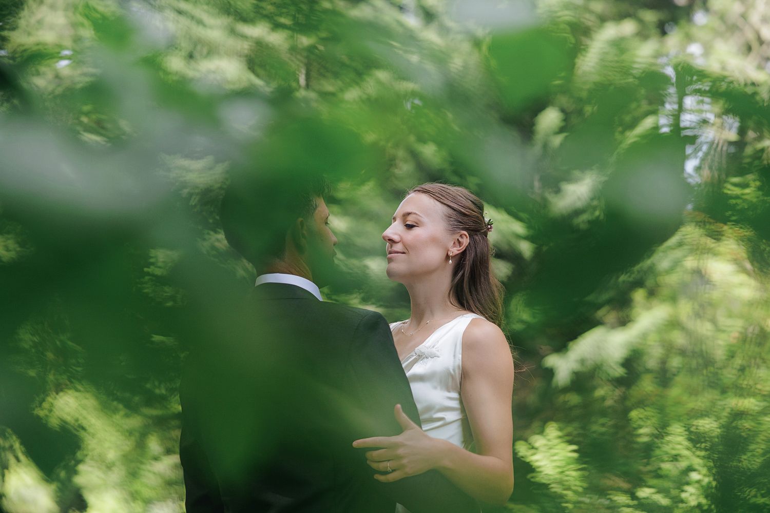 A dreamy summer wedding scene through green foliage, with a bride in a white sleeveless dress captured in soft focus.