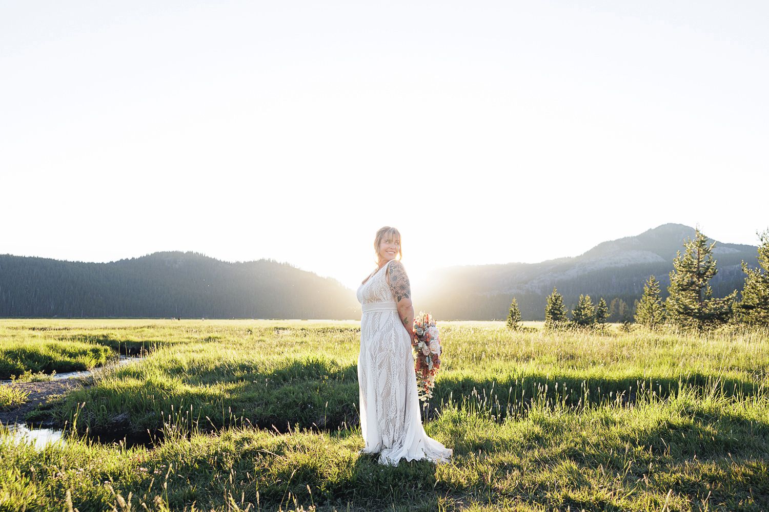 A person in a flowing white dress stands in a sunlit mountain meadow at golden hour.