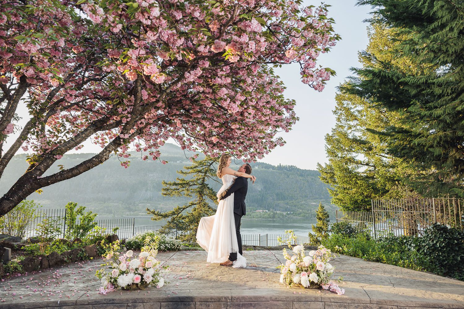 A couple shares a romantic moment under blooming cherry blossoms overlooking a mountain lake with floral decorations.