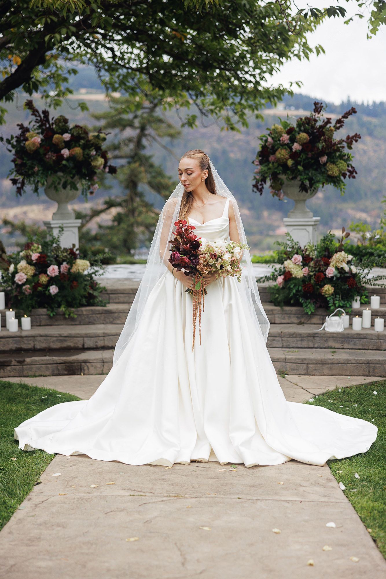 Bride in white gown and veil holds bouquet at outdoor ceremony with floral arrangements and mountain backdrop.